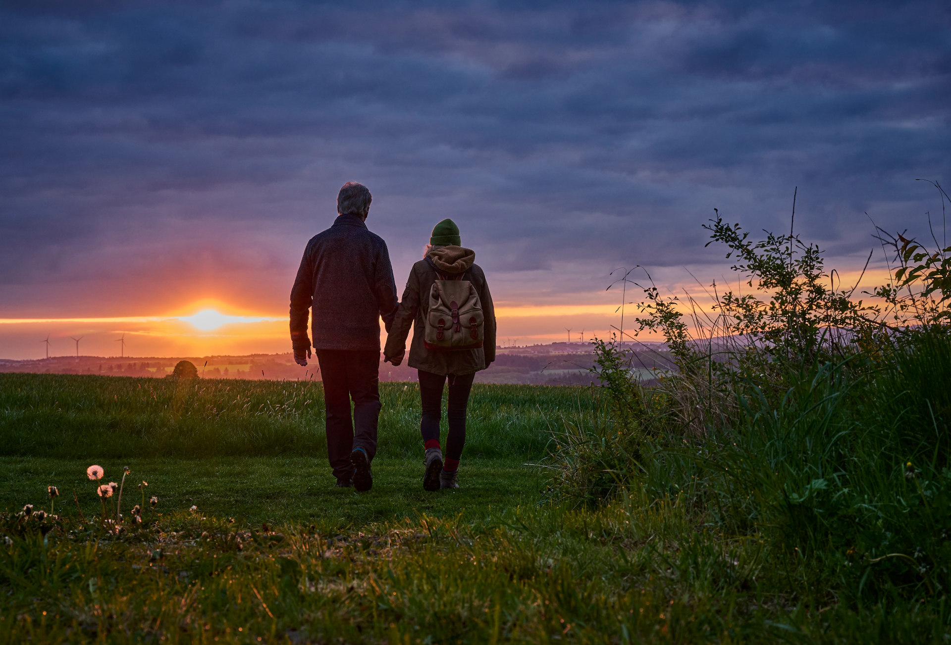 Zwei Personen, die sich beim Wandern den Sonnenuntergang im Hunsrück mit Blick auf Wäldern, Windräder und das Umland anschauen