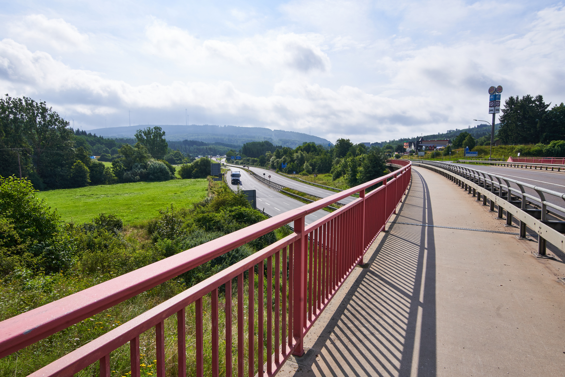 Blick von einer Fuß- und Radwegbrücke mit rotem Geländer auf eine Bundesstraße, umgeben von grünen Wiesen, Bäume und hügeliger Landschaft im Hunsrück.