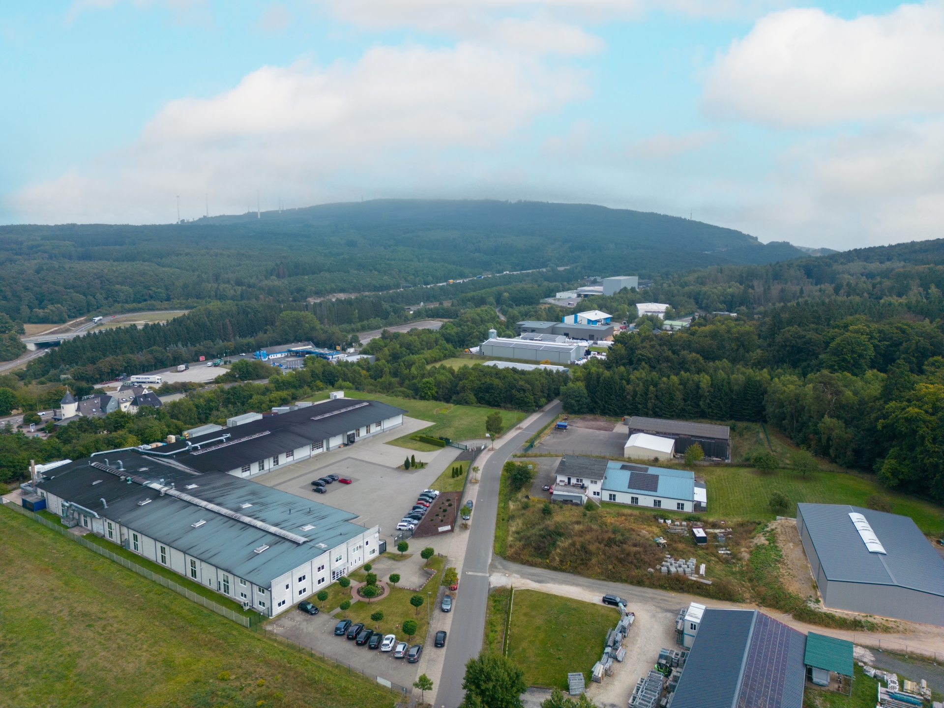Industriegebiet und Gewerbegebiet von Rheinböllen mit Produktionshallen, Lagerhallen und Büros, umgeben von bewaldeten Hügeln und einer Landstraße, unter bewölktem Himmel.